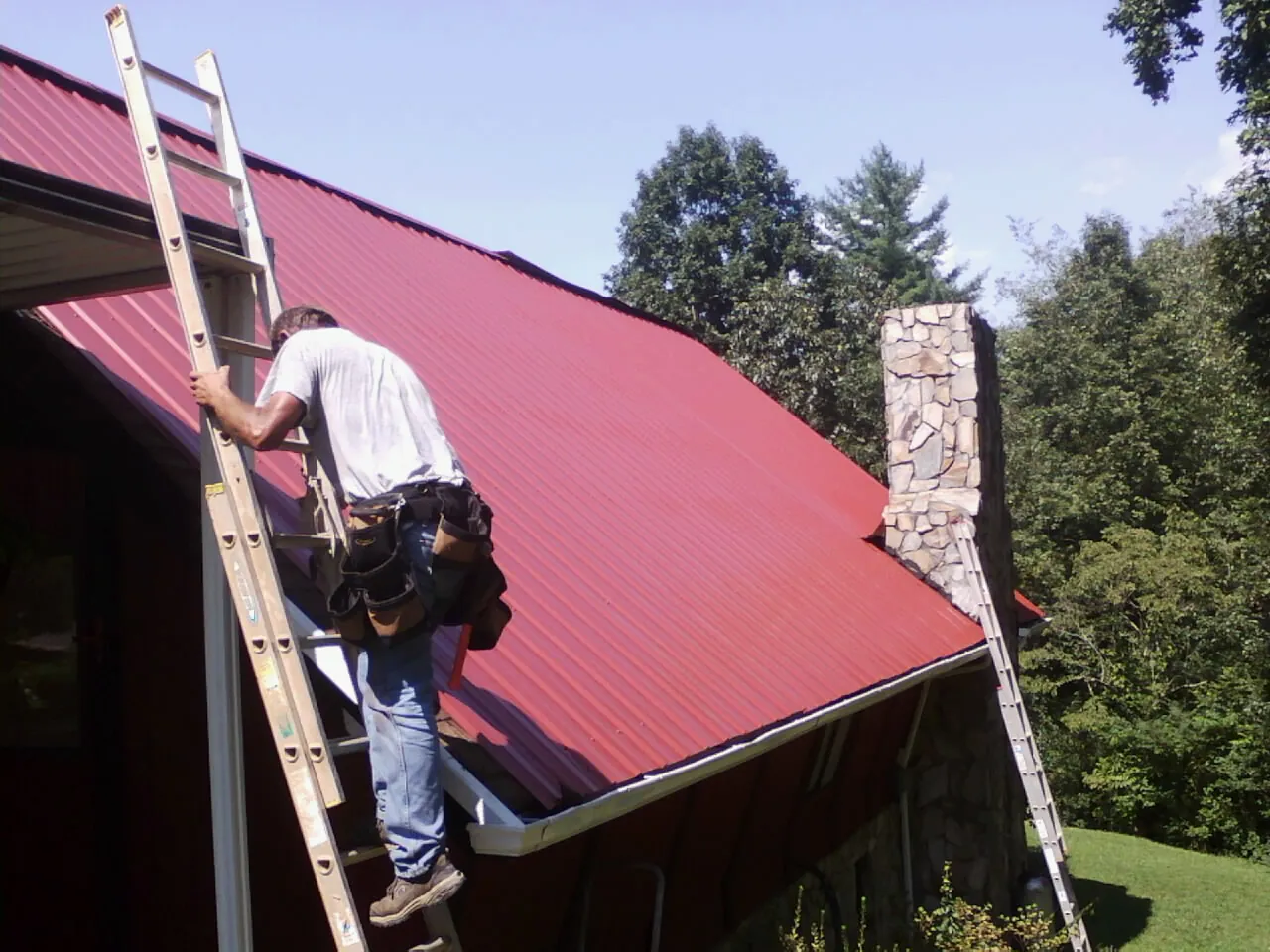 Roofer on a ladder beside a red metal roof and stone chimney — Soffit & Fascia Repair by Roofing Brothers in Rapelje